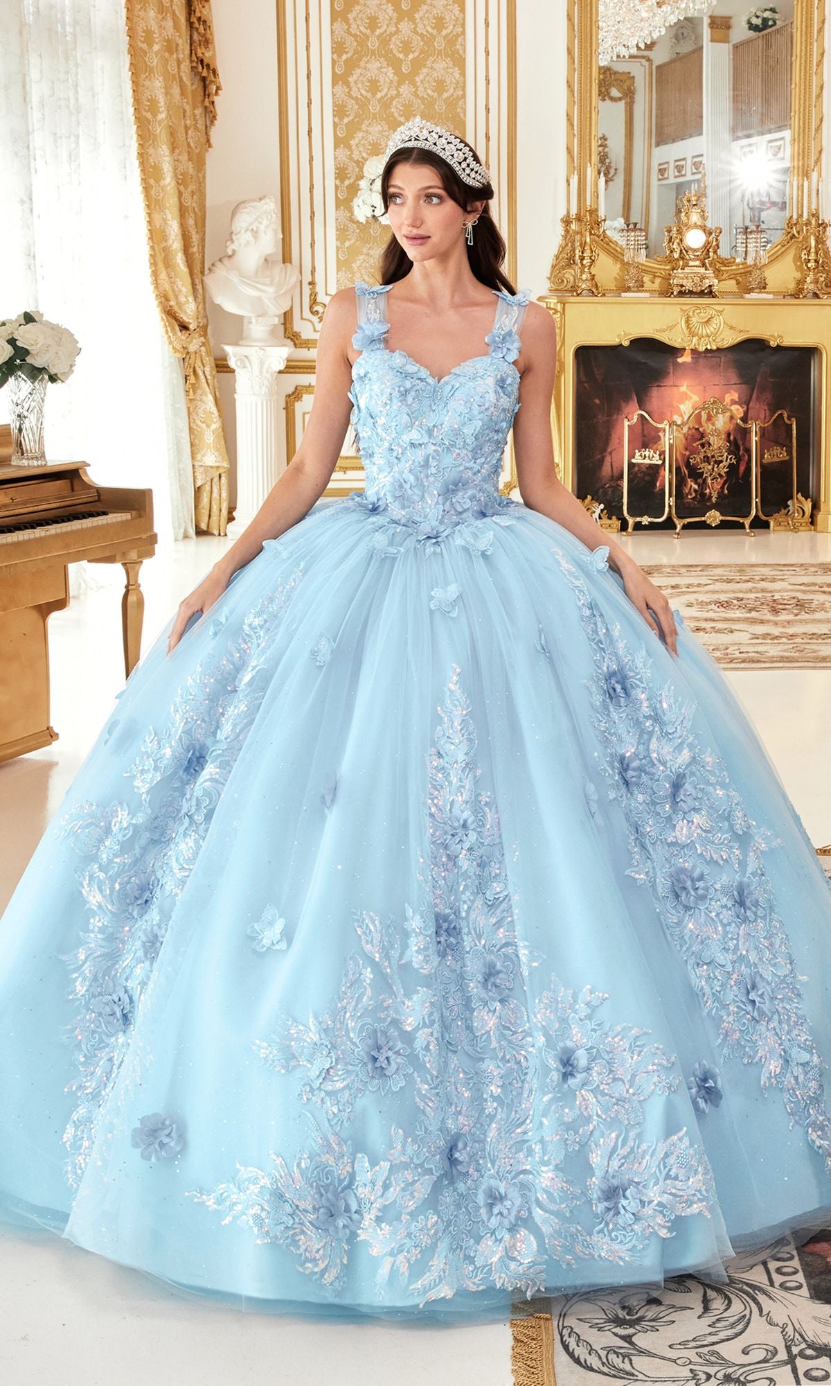 A woman stands indoors in an ornately decorated, gold-accented room, wearing the Quinceanera Dress 15713 by Ladivine—an off-the-shoulder light blue gown with floral embroidery and a jeweled headband.